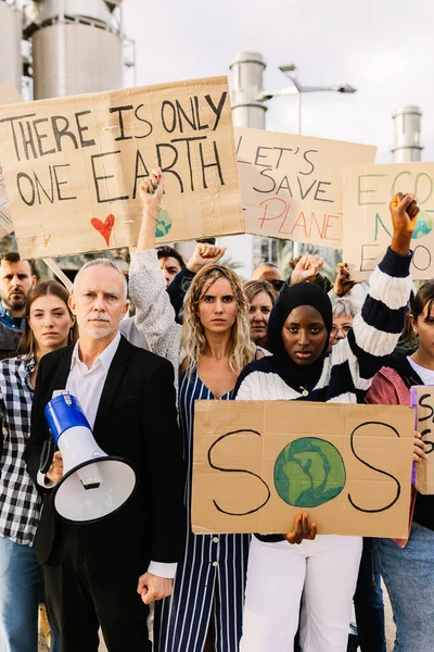 Organized group of diverse people with posters and placards protesting against climate change in the world in front of a polluting factory. Environmental conservation and global warming concept.