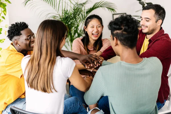Diverse group of people gathered at therapy session. Millennial people sitting on chairs in circle stacking hands supporting each other at community center. Help and support concept.