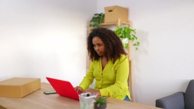 Genç Afrikalı Amerikalı küçük işletme sahibi kadın ofiste çalışırken dizüstü bilgisayar kullanıyor. Kadın işçi atölyede yeni bir teslimat hazırlıyor. E-ticaret ve girişimci insan kavramı.