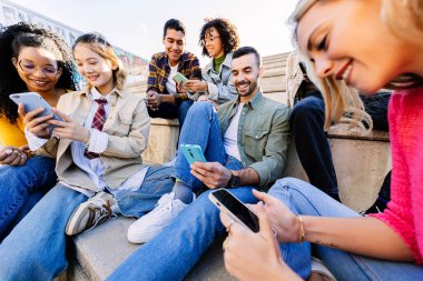 Açık havada yan yana oturarak cep telefonu kullanan bağımlı gençler. Milenyum arkadaş grubu cep telefonu ekranına bakıyor, video oyunları oynuyor veya uygulamadan sosyal medya içeriğinden keyif alıyor.
