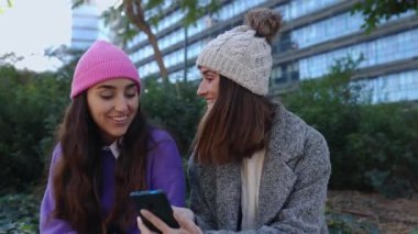 Akıllı telefon ekranına bakan iki genç kadın ağ uygulamasında sosyal medya içeriğinin keyfini çıkarıyorlar. Kışın dışarıda oturan kız arkadaşlar cep telefonlarını birlikte kullanıyorlar..