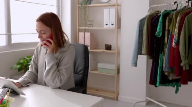 Atölyede çalışırken cep telefonuyla konuşan neşeli beyaz bir kadın. Küçük işletme sahibi kadın dijital tablet kullanarak internette geziniyor. E-ticaret ve iş konsepti.