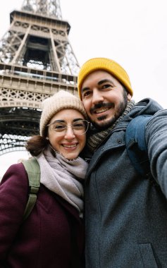 Paris, Fransa 'daki Eyfel Kulesi' nin önünde telefonla selfie çeken genç Avrupalı bir çiftin dikey fotoğrafı. Seyahat ve tatil kavramı.