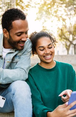 İki genç Latin Amerikalı 'nın dışarıda otururken akıllı telefon kullanışının dikey görüntüsü. Neşeli Afrikalı Amerikalı arkadaşlar cep telefonu ekranına bakarken gülüyorlar. Sosyal medya kavramı.