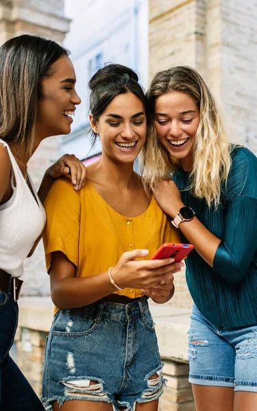 Multiracial young female friends laughing while using mobile phone outdoors. Three diverse women having fun watching funny content on smart mobile phone device.