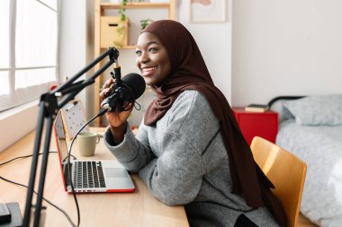 Genç Afrikalı Müslüman kadın odasında dizüstü bilgisayarı ve mikrofonuyla podcast yapıyor.