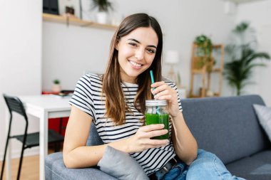 Kanepede dinlenirken elinde yeşil detoks suyu tutan mutlu genç bir kadın. Sağlıklı yaşam tarzı ve diyet kavramı.