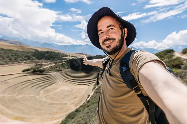 Happy young tourist man taking selfie portrait in Moray, agricultural terraces in Peru. Joyful tourist enjoying summer vacation in South America.