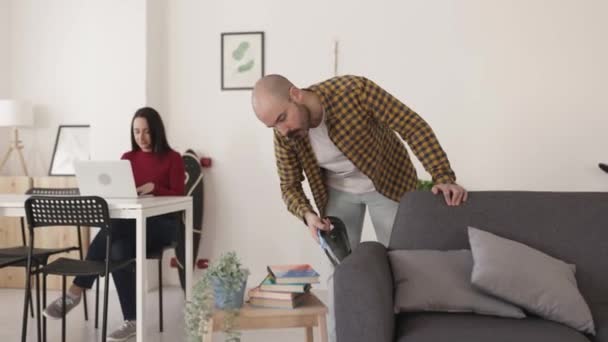 Hipster Homem Adulto Jovem Limpando Sofá Com Aspirador Enquanto Mulher — Vídeo de Stock