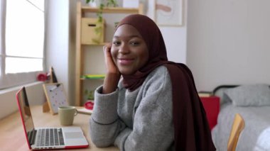 Masada oturan ve kameraya gülümseyen Afrikalı yaratıcı bir kadının portresi. Müslüman başörtüsü takmış mutlu siyah kız öğrenci evde dinleniyor. Gençlik ve teknoloji kavramı. Yüksek kalite 4k görüntü