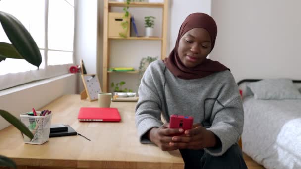 イスラム教徒のヘッドスカーフの幸せな遺伝子Zの若い黒人女性は 家庭で携帯電話を使用しています ソーシャルメディア 若者のライフスタイル テクノロジーコンセプト — ストック動画