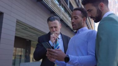 Şehir caddesinde akıllı telefon kullanan başarılı bir grup iş adamı. Üç nesildir cep telefonu ekranına bakan iş adamı ofis binasının önünde yan yana duruyor..