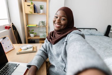 Müslüman başörtüsü takmış neşeli siyah Afrikalı kadın yatak odasında telefonuyla selfie portresi çekiyor..
