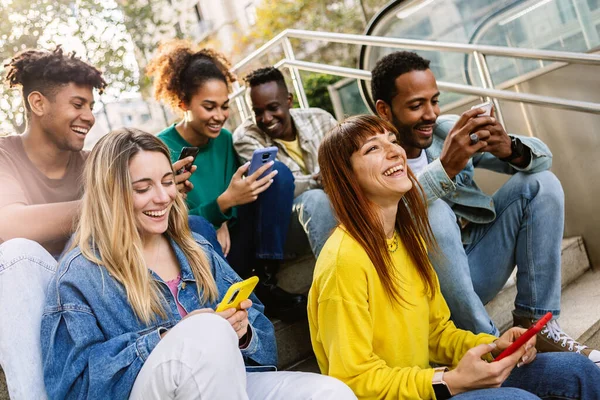 Young group of happy people using mobile phone relaxing together outdoors. Smiling multiracial friends sitting at the subway entrance looking social media content on cellphone. Youth lifestyle.