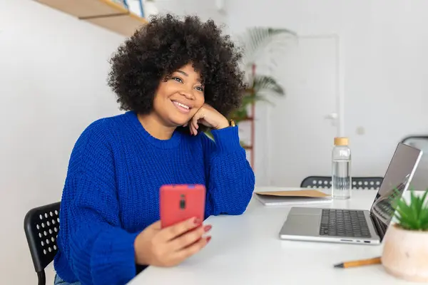 Happy beautiful young african woman holding mobile phone working on laptop at home. Technology concept