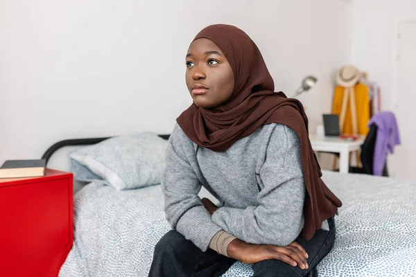Sad young african woman in muslim headscarf sitting on bed looking through the window at home