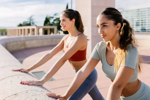 Side view of two young fit caucasian women wearing sports clothes stretching in an urban park. Active lifestyle and sport concept.