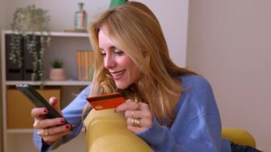 Evde cep telefonu uygulaması kullanarak internetten alışveriş yaparken kredi kartı tutan mutlu genç sarışın kadın. E-ticaret ve teslimat hizmeti kavramı.
