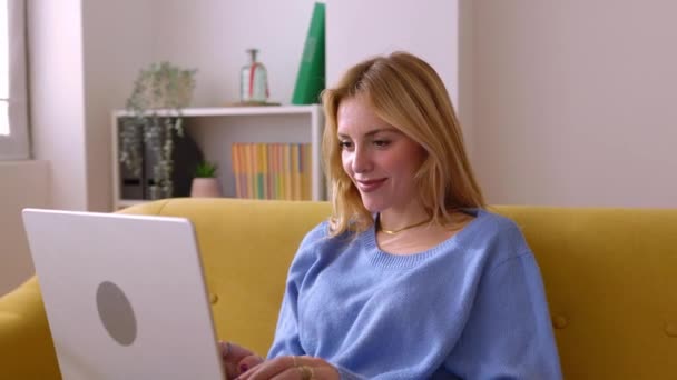 快乐的年轻金发女人坐在沙发上 一边用笔记本电脑 一边坐在家里 千年女学生在家网上学习 — 图库视频影像