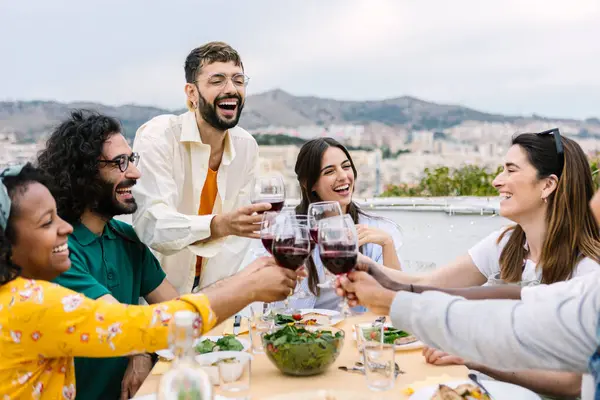 Group of young friends having dinner at home terrace. Millennial people enjoying time together sitting on table at summer party celebration. Friendship concept