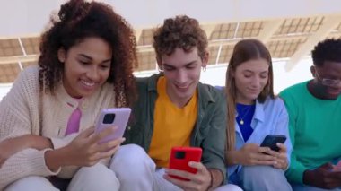 Genç bir grup çok ırklı arkadaş dışarıda birlikte oturan cep telefonu cihazını kullanarak eğleniyor. Milenyum insanları cep telefonu uygulamasında sosyal medyadan keyif alıyorlar. Gençlik kavramı.