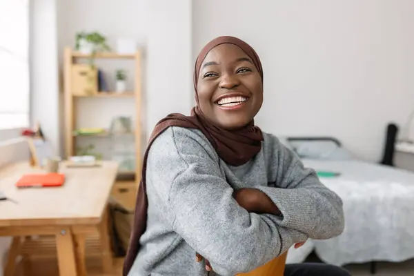 Joyful portrait of young african girl relaxing on her room. Positive gen z black female sitting on chair indoors. Happiness and proud people concept.