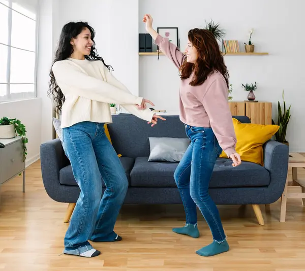Two young diverse female friends having fun dancing in the living room at home. Friendship and domestic lifestyle concept.