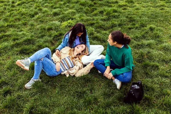 Three young diverse girls relaxing together at city park. Female friends lying on college campus grass, talking and enjoying time.