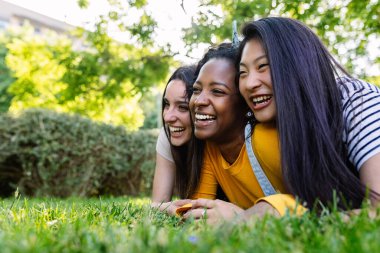 Üç genç milenyum kadını şehir parkında çimlere uzanıp gülüyor, yazın birlikte vakit geçiriyorlar..