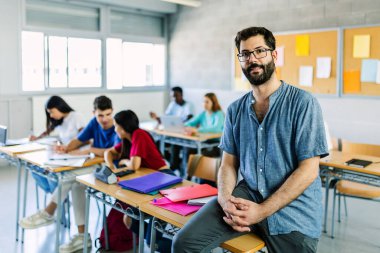 Portrait of young bearded male teacher looking at camera while sitting at high school table over group of diverse students learning together. Education lifestyle concept clipart