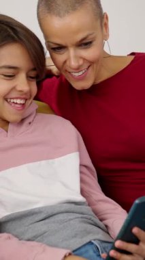 Bekar bir anne ve kızı koltukta dinlenirken cep telefonundan komik videolar izliyorlar. Teknoloji ve ev hayatı kavramı. Dikey HD video görüntüleri.