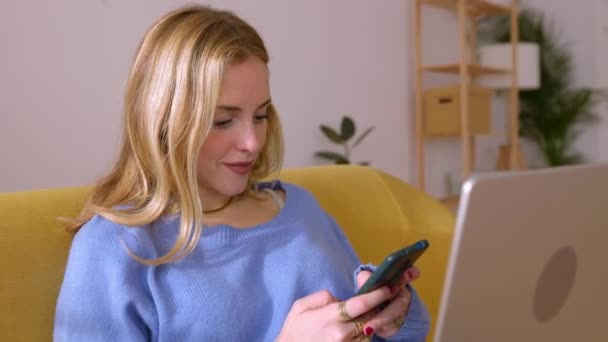 自宅でソファーに座っている間 携帯電話を使用してラップトップを持つ幸せな若い女性 技術ライフスタイルコンセプト — ストック動画