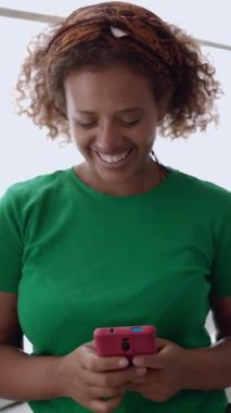 Genç Afrikalı kadın cep telefonu uygulamasında sohbet ederken eğleniyor. Evde akıllı telefon kullanarak gülümseyen melez bir kadın. Dikey HD ekran görüntüleri.