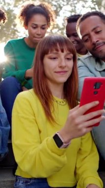 Dikey HD ekran video. Genç ve mutlu arkadaşlar cep telefonlarıyla dışarıda oturarak eğleniyorlar. Milenyum insanları akıllı telefon uygulamasında komik sosyal medya içeriğini izlerken gülüyorlar.