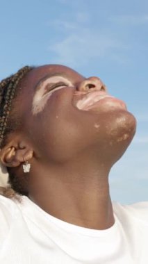 Dikey video. Kapalı gözlü Afrikalı genç bir kız dışarıda durup güneşe dönüşmüş güneşin tadını çıkarıyor, sıcak bir günün tadını çıkarıyor ve özgür hissediyor. Refah ve özgürlük kavramı.