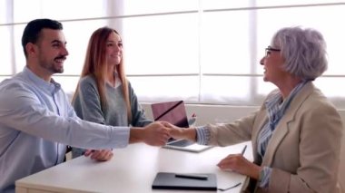 Mutlu gülümseyen milenyum çifti, anlaşma ya da sözleşme imzaladıktan sonra ofiste üst düzey kadın emlakçıyla tokalaşıyor..