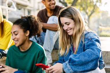 Mutlu öğrenci arkadaşlar dışarıda otururken birlikte cep telefonu kullanıyorlar. Milenyum çeşitliliği olan insanlar cep telefonlarıyla konuşuyor. Şehir caddesinde dinleniyorlar. Gençlik yaşam tarzı konsepti.