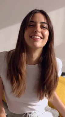Genç ve mutlu bir kadının sabahleyin odasında yatağında kameraya gülümseyişini gösteren dikey bir video. Yatak odasında dinlenen güzel bir kadının portresi.. 