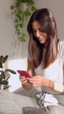 Akıllı telefon kullanan mutlu genç bir kadının sabahları yatakta oturduğu dikey bir video. Milenyum kadını cep telefonu uygulamasındaki sosyal medya içeriğini izleyerek eğleniyor. Hafta sonları odada dinleniyor..