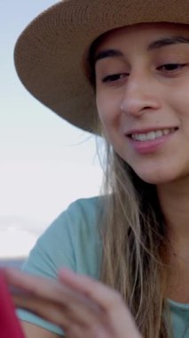 Yazlık şapkalı, dışarıda cep telefonu kullanan, internette sörf yapan ya da sosyal ağ uygulamasında medya içeriğini izleyen mutlu bir genç kadın. Teknoloji yaşam tarzı konsepti. Dikey HD ekran videosu