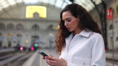 Tren istasyonunda tren beklerken cep telefonu kullanan mutlu genç bir gezgin kadın. Banliyö insanları ve teknoloji yaşam tarzı konsepti.