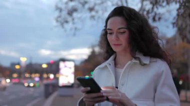 Arka planda şehir ışıkları olan caddede akıllı telefon kullanan mutlu genç bir kadın.