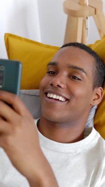 Dikey HD video. Kanepede cep telefonu kullanan genç bir adam. Milenyum erkeği akıllı telefon uygulamasında evde gevşeyerek sohbet ediyor. Teknoloji yaşam tarzı konsepti.