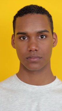 Dikey HD video. Beyaz tişörtlü Afro-Amerikan genç adamın yakın plan portresi. Sarı arka planda kameraya ciddi ciddi bakıyor. Gençlik ve Gen Z İnsanlar konsepti.