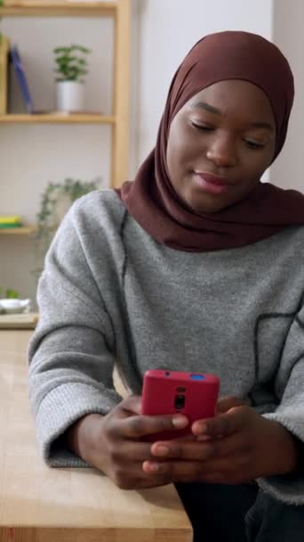 イスラム教徒のヘッドスカーフの ハッピー ジェンツの若いアフリカ人女性が 自宅で携帯電話を使っています ソーシャルメディア 若者のライフスタイル テクノロジーコンセプト 垂直Hd ビデオ — ストック動画