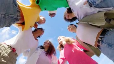 Çemberde bir araya gelip birbirlerine sarılmış ve kameraya gülümseyen çeşitli genç kadınların düşük açılı görüntüsü. Kadın arkadaşlığı ve toplum, feminizm ve birlik kavramı.