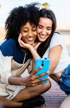 Genç, mutlu, çok ırklı kadınların yaz aylarında açık havada akıllı telefon uygulamalarıyla selfie çekerken çekilmiş dikey fotoğrafı. LGBT lezbiyen çifti fotoğraf çekiyor. Kadın arkadaşlığı kavramı. 