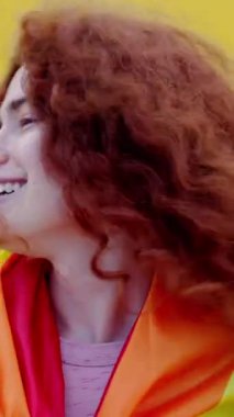 Sarı arka planda kameraya gülümseyen gökkuşağı bayrağı taşıyan kızıl saçlı, ikili olmayan adamın dikey portresi. LGBT topluluğu, çeşitlilik, cinsiyet değiştirme ve ikili olmayan insan kavramı