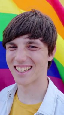 Sarı arkaplanda kameraya gülümseyen genç adamın dikey portresi elinde kameraya gülümseyen gökkuşağı bayrağı. LGBT topluluk kavramı.