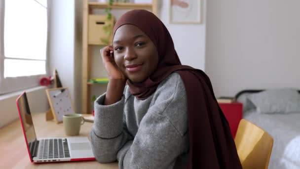 一个年轻的非洲创意女人坐在办公桌前对着相机微笑的画像 穿着穆斯林头巾的快乐的黑人女生在家里放松一下 青年与技术概念 高质量的4K镜头 — 图库视频影像